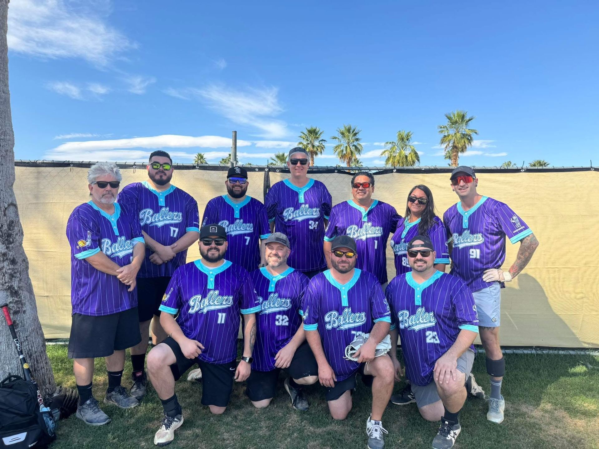 Group of people in purple jerseys posing outdoors in front of a wall and palm trees.