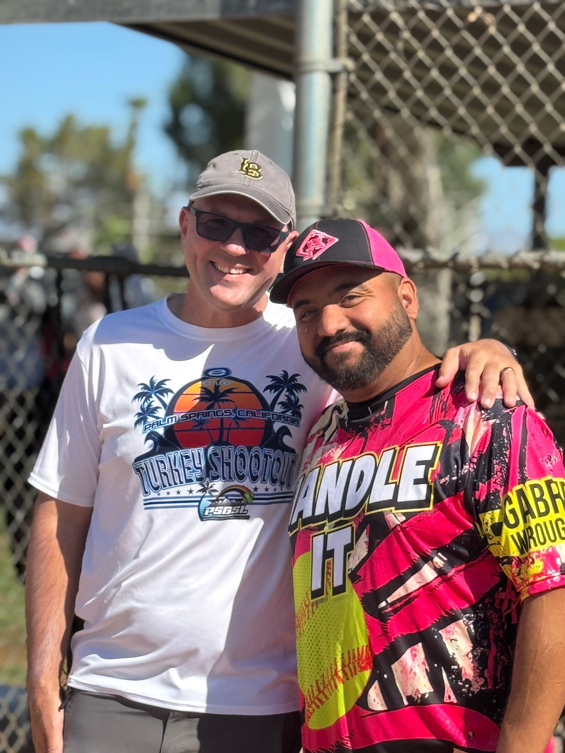 Two men smiling outdoors, one in a white shirt and cap, the other in a pink jersey and cap, with an arm around his shoulder