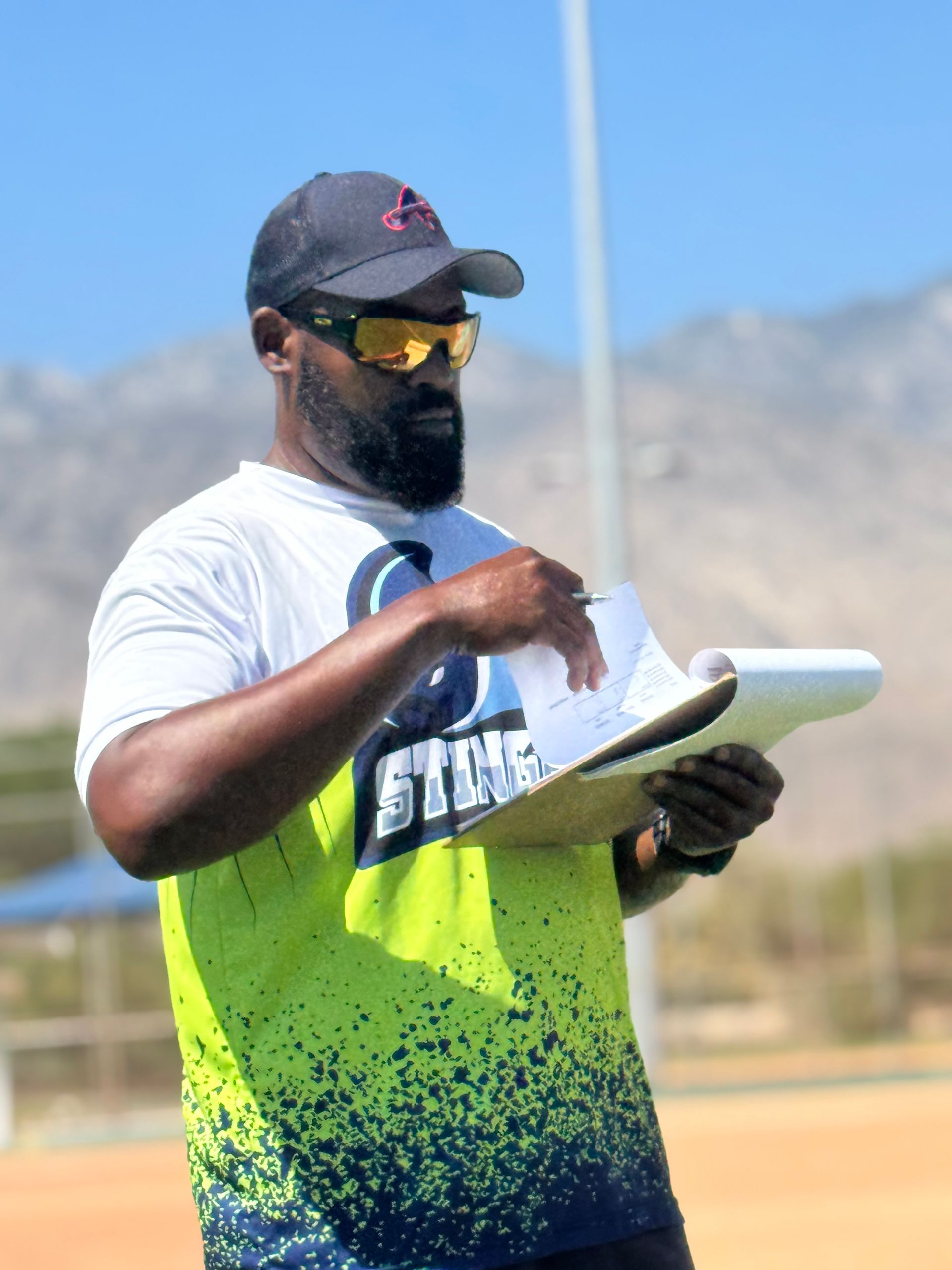 Man in sunglasses and cap holding a clipboard outdoors with mountains in the background