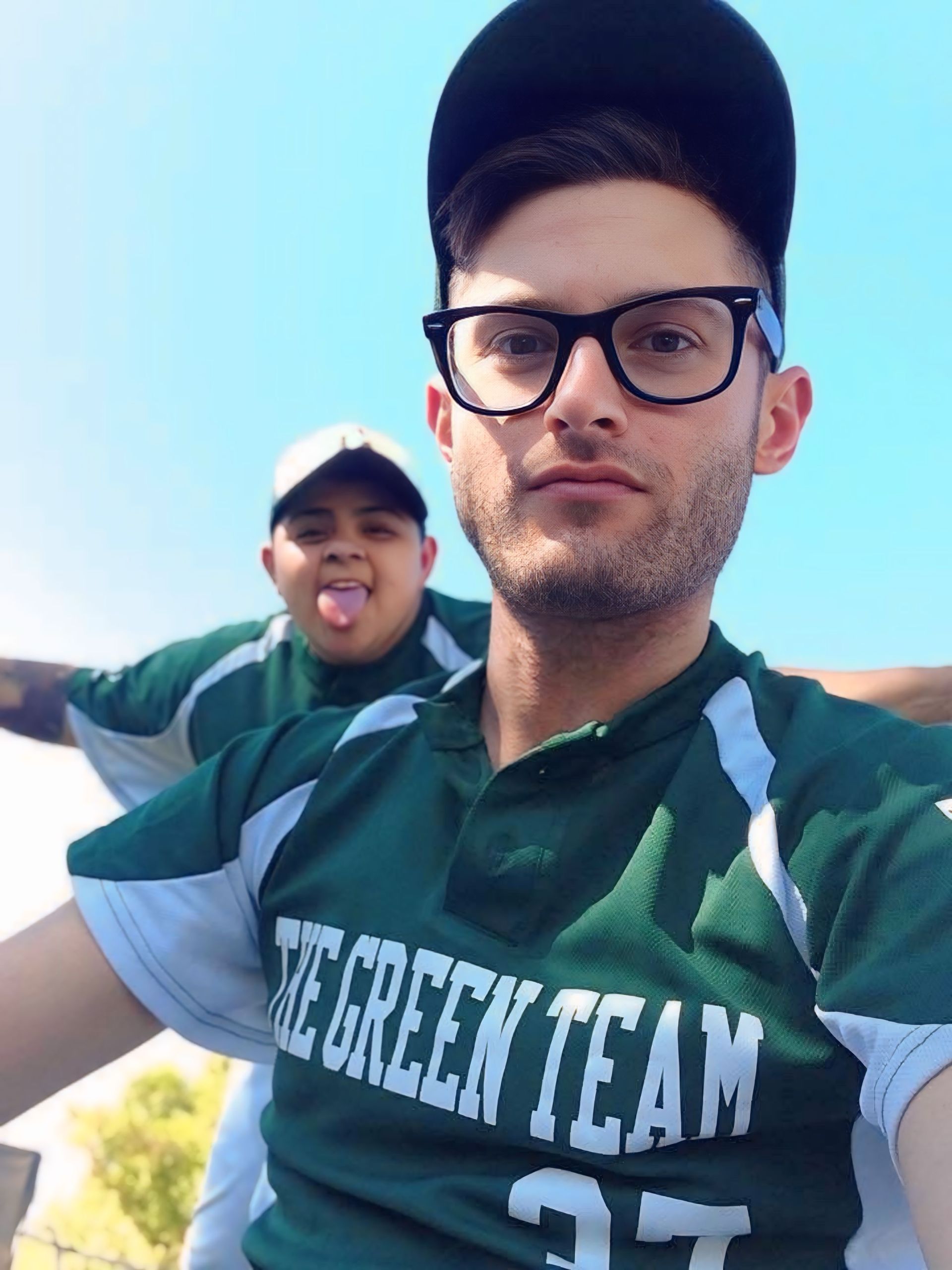 Two fans in green jerseys at a game, one in front with glasses and cap, one behind playfully sticking out tongue