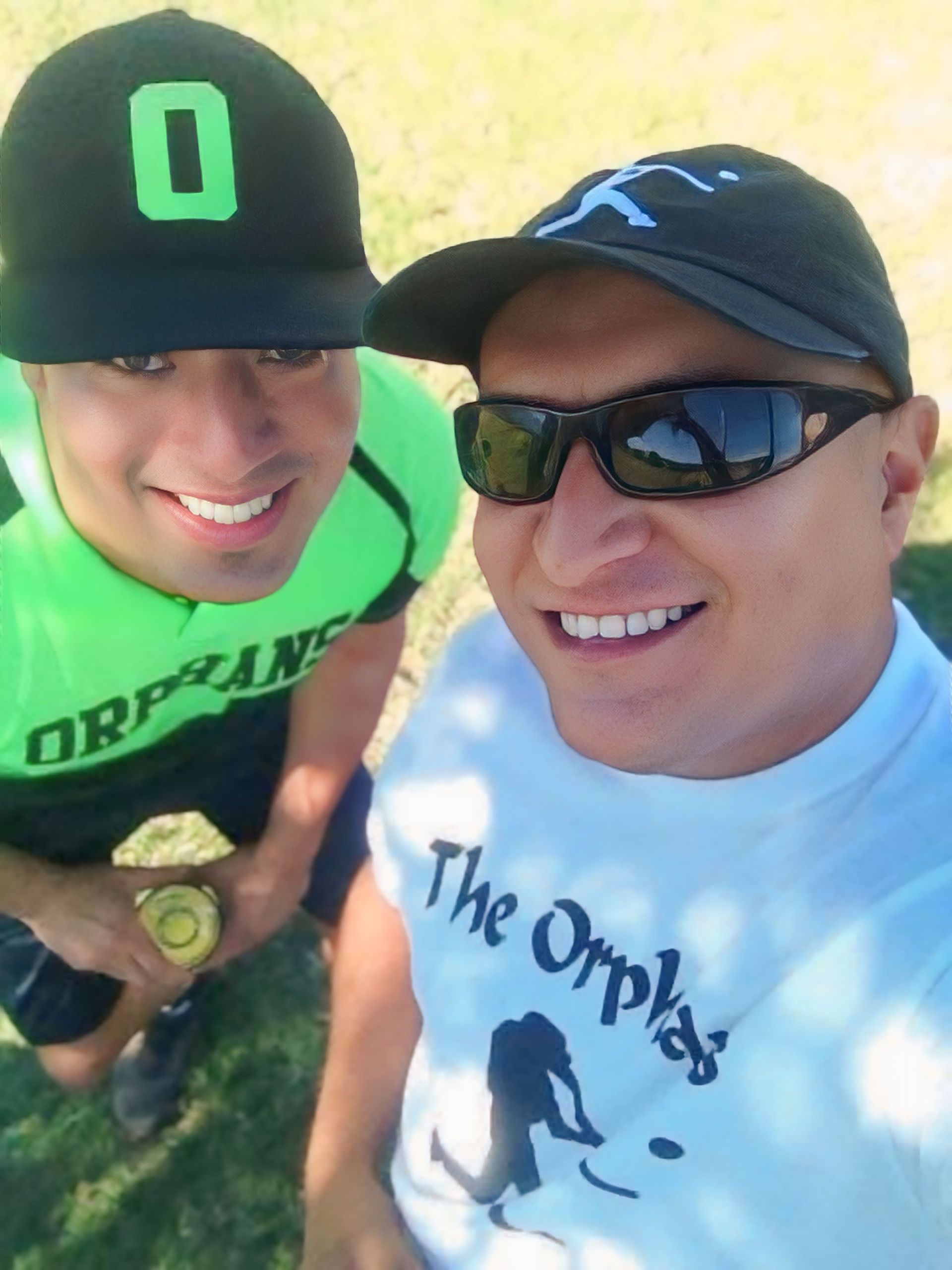 Two smiling people posing outdoors in hats and sunglasses, one in a green hoodie and one in a white T-shirt.