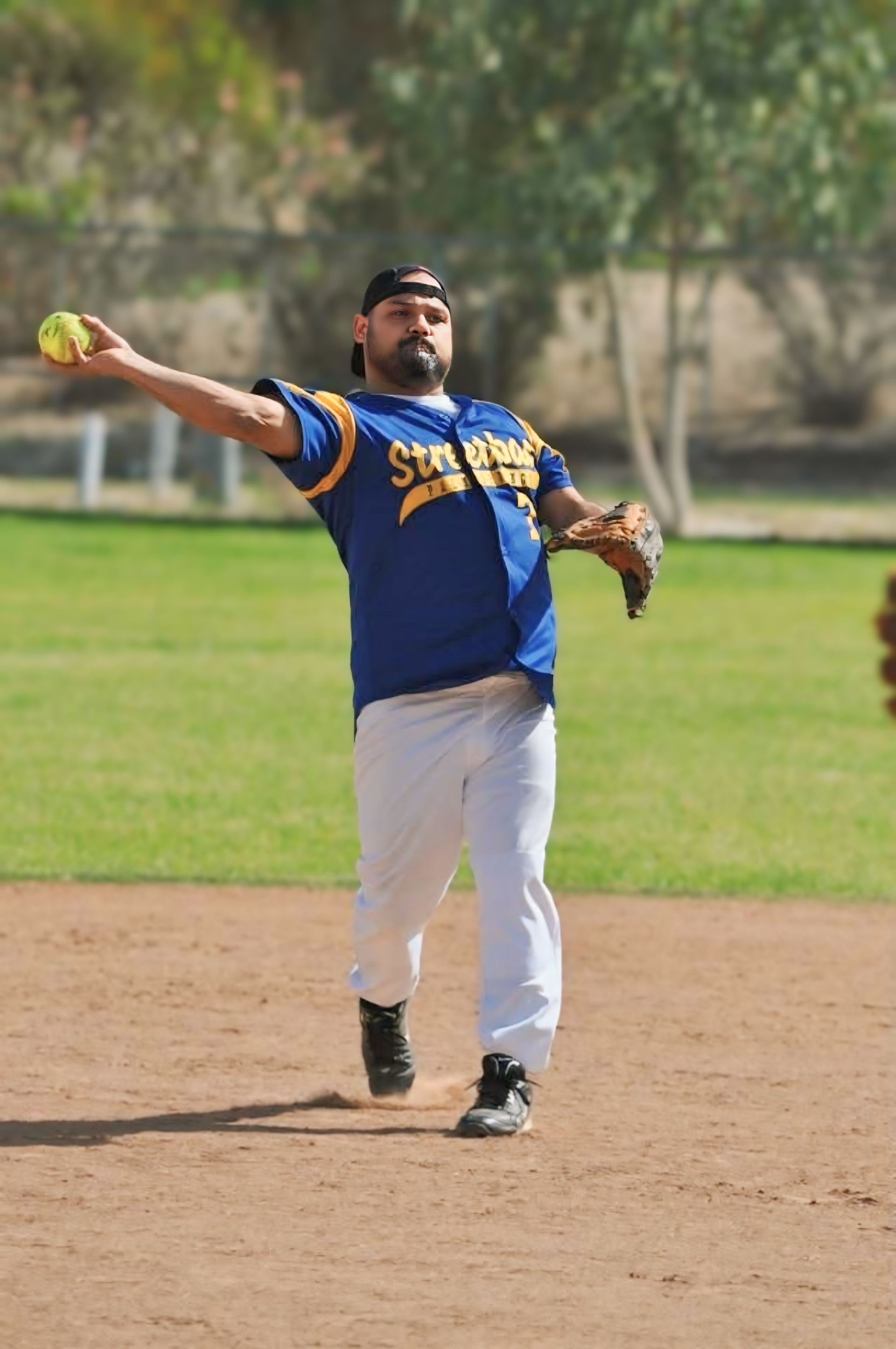 Softball pitcher in blue and yellow uniform throwing a ball on a dirt field