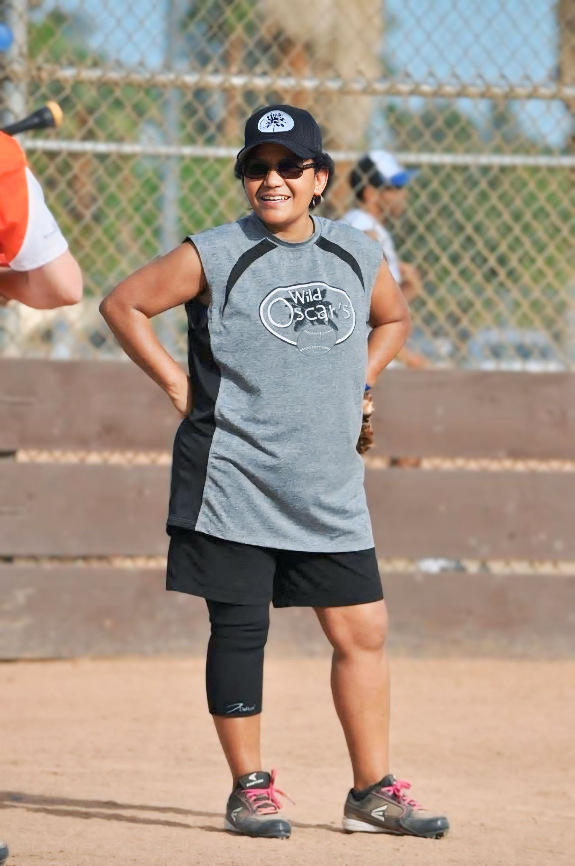 Woman in gray athletic shirt and black shorts stands on a softball field, hands on hips, wearing a cap and sunglasses