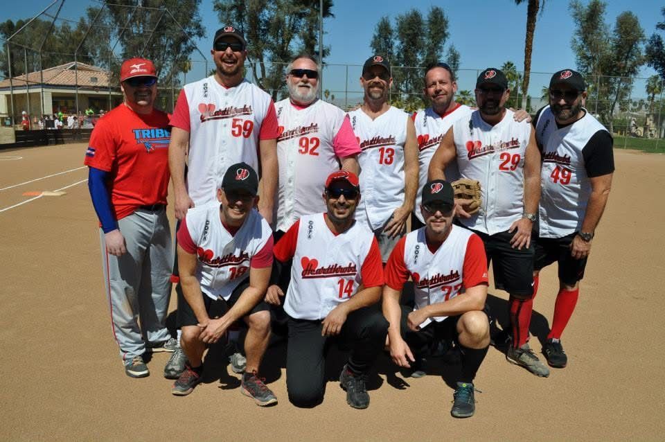 Softball team posing outdoors on a sunny field, wearing red-and-white jerseys and caps, with a blue sky behind them