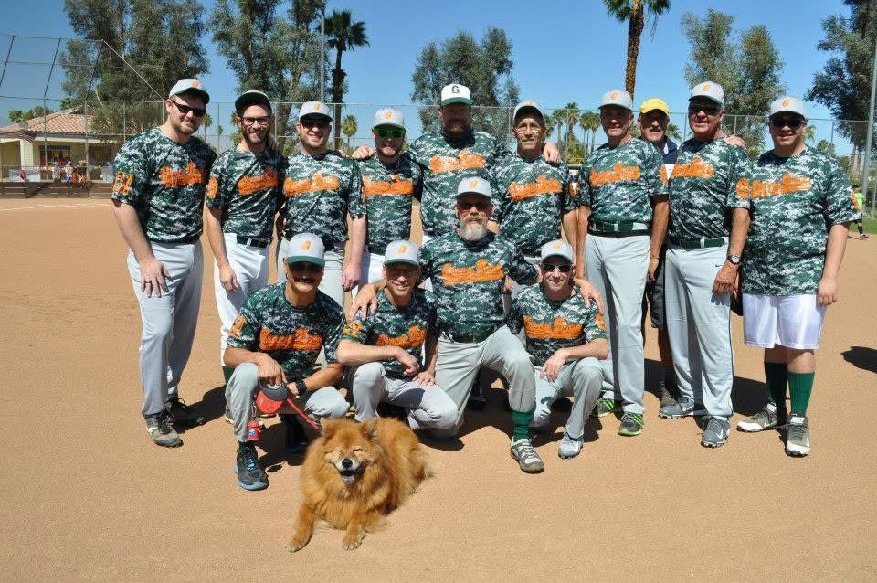 Group of baseball players in camo jerseys posing on a sunny field with a golden retriever in front