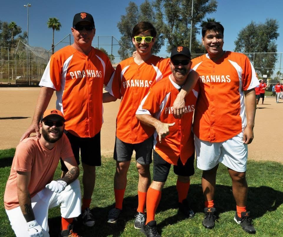 Five people in orange and white sports uniforms posing on a baseball field under a sunny sky