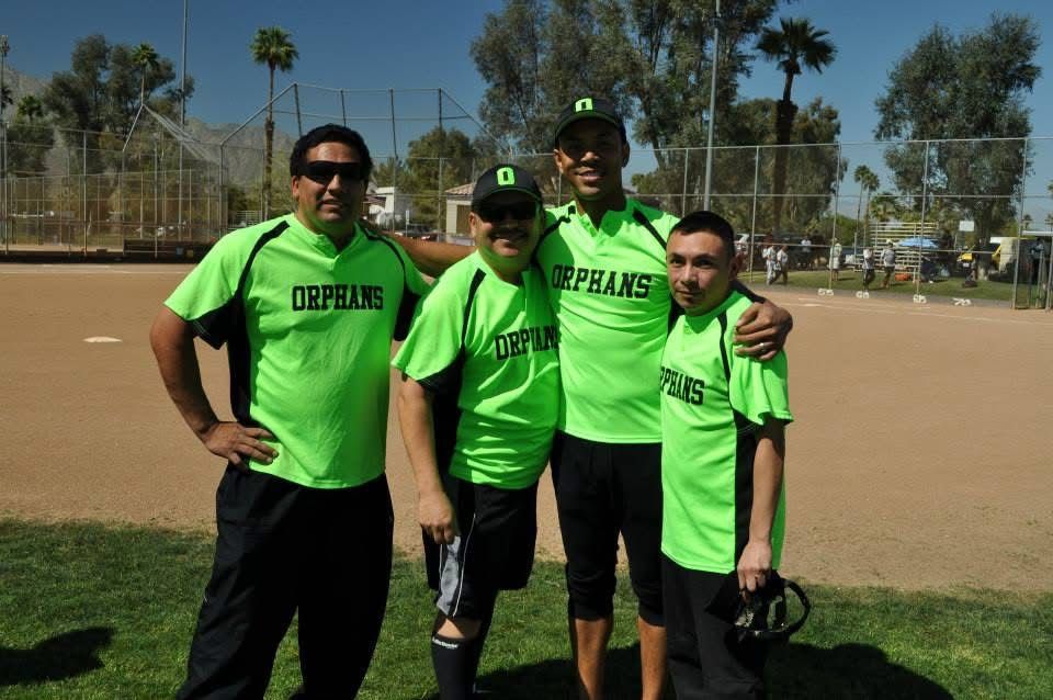 Four teammates in neon green shirts pose on a baseball field, holding gloves under a sunny sky