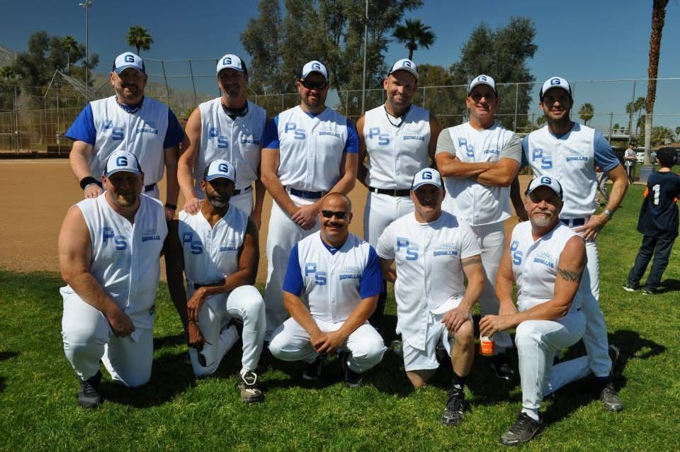 Sports team posing outdoors on grass, wearing white uniforms and caps, with palms and blue sky in the background