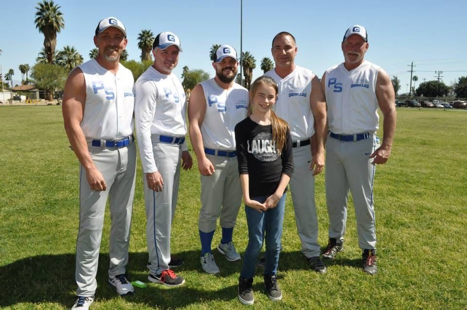 Five baseball players in white and gray uniforms standing with a girl on a grassy field under a blue sky