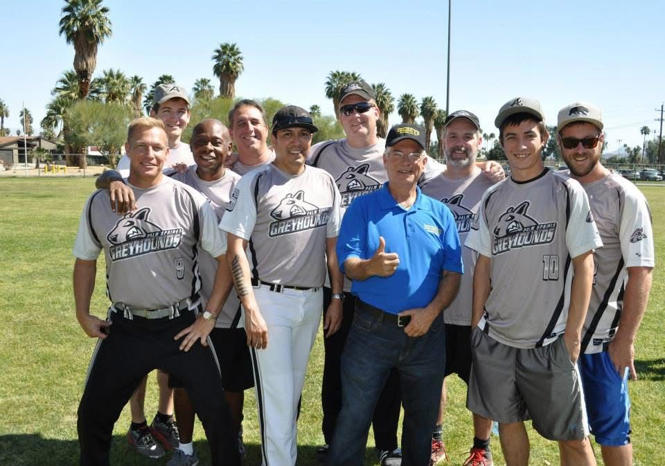 Group of men posing on a sunny sports field, several in matching gray shirts and one in blue.