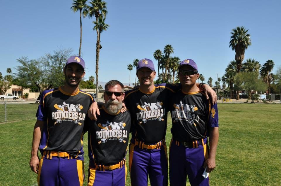 Four softball players in black and purple uniforms standing together on a sunny field