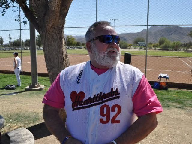 Man in pink jersey and sunglasses standing on a baseball field near a tree, with mountains in the background