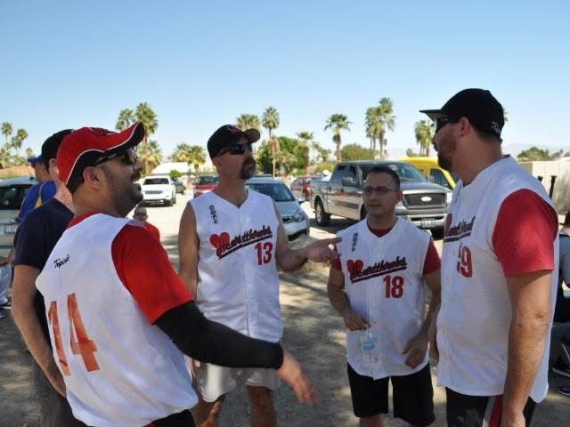 Four softball players in white-and-red jerseys talking together outdoors near parked cars.