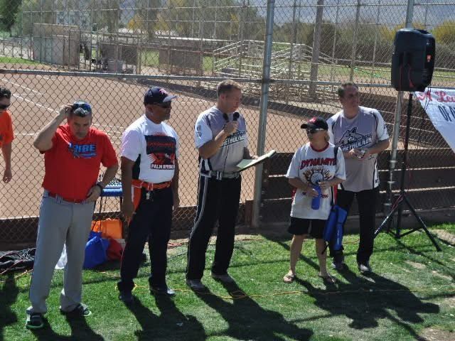 Five people standing by a chain-link fence at a sports field, holding items and smiling at the camera