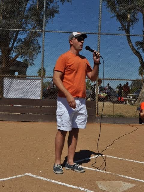 Man speaking into a microphone on a tennis court, wearing an orange shirt and white shorts.
