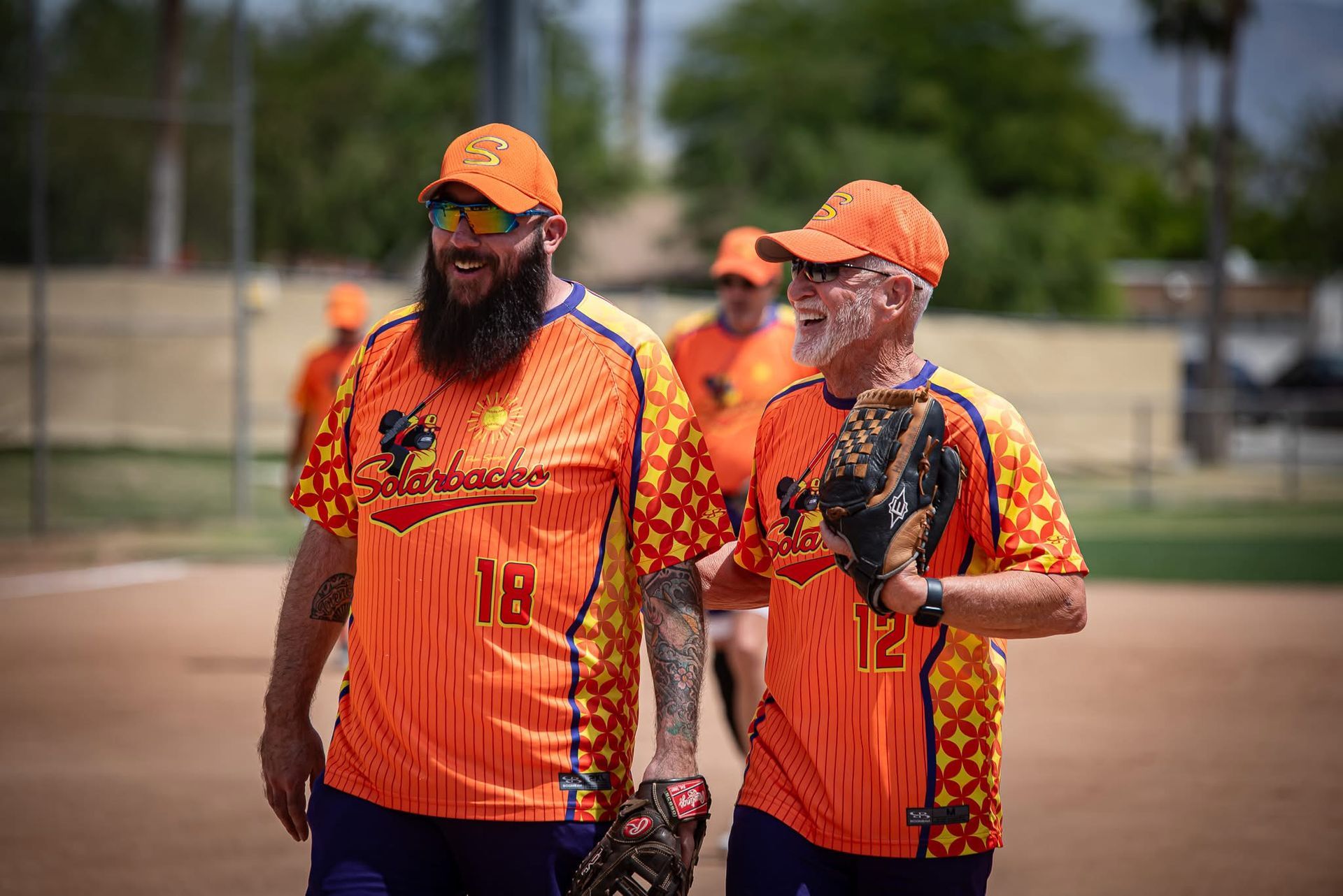 Two softball coaches in orange jerseys and caps stand on the field, talking before a game.