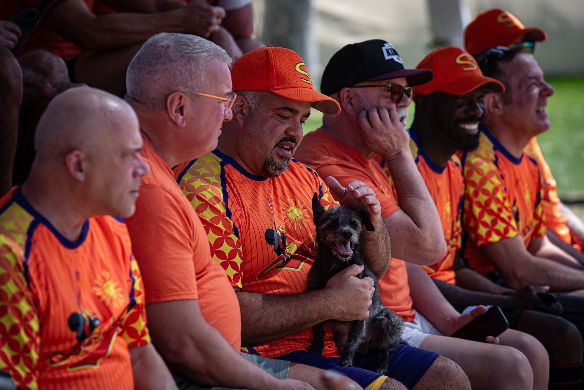 Row of fans in orange shirts and caps sitting together in stadium seats, one holding a small dog