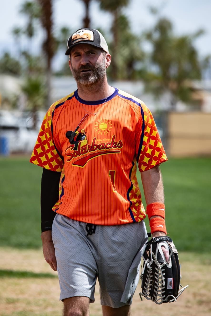 Baseball player in orange jersey and gray shorts walking on the field, holding a glove and ball cap.