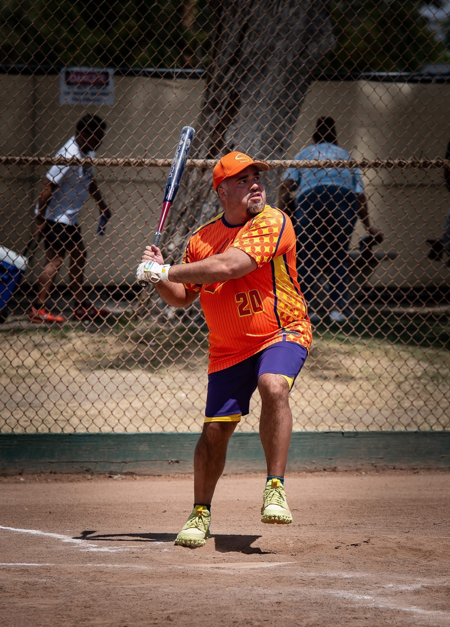 Softball batter in orange shirt and cap swings at a pitch on an outdoor field.
