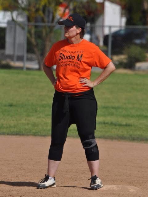 Softball player in orange jersey and black pants standing on a field with hands on hips.