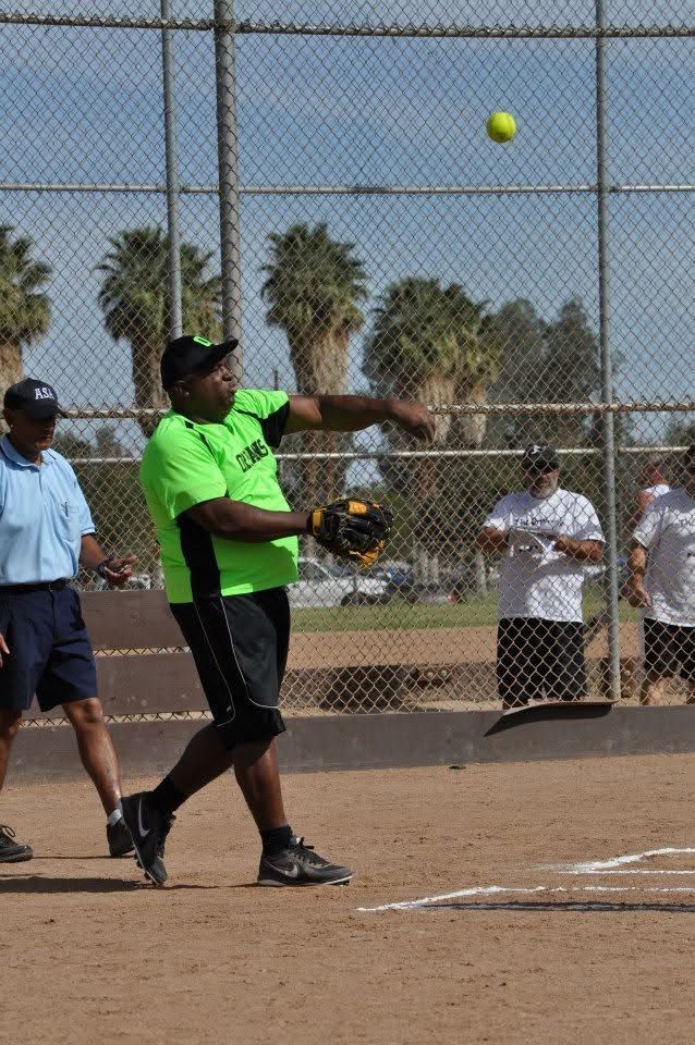 Softball player in neon green shirt swings at a pitch on a dusty field with chain-link backstop.