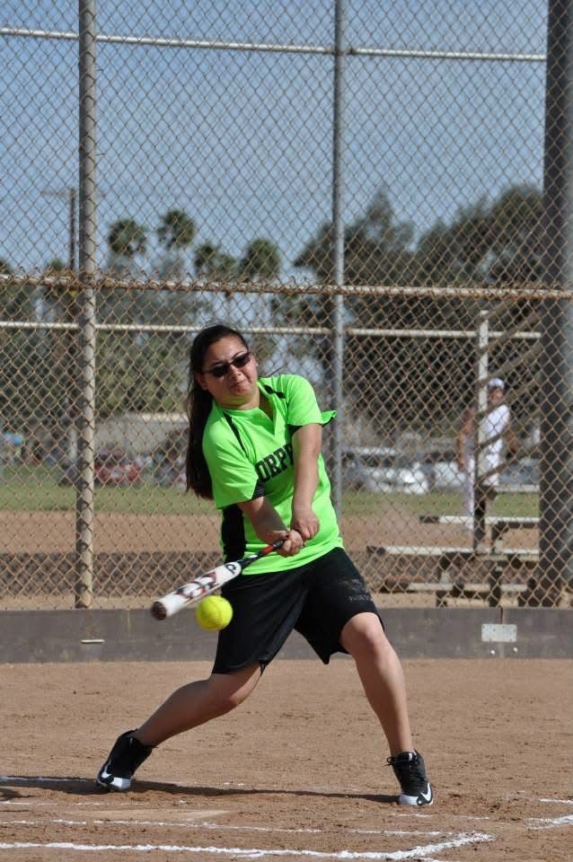 Softball player in neon green shirt swings at a pitch on a dirt field, with chain-link fencing behind her
