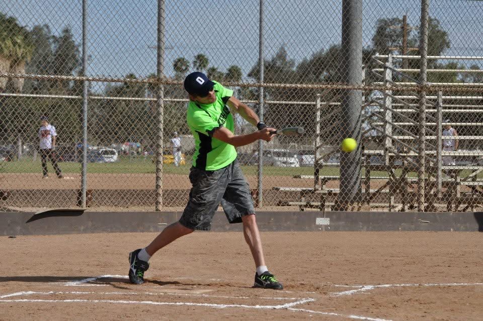 Softball player in neon green swings at a pitch on a dirt field, with chain-link backstop behind him