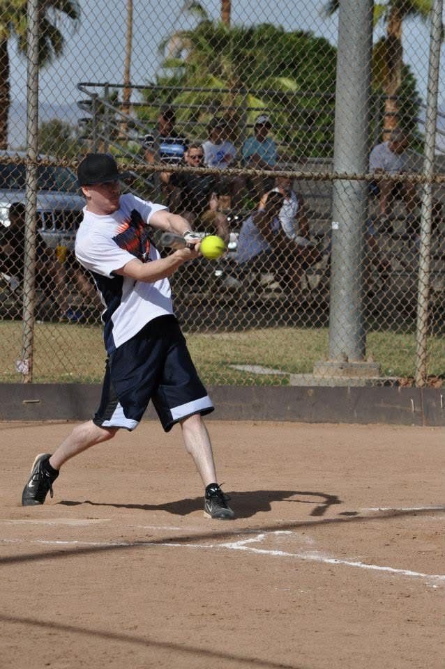 Baseball player in white and black swings at a pitch on a dirt field behind a chain-link fence.