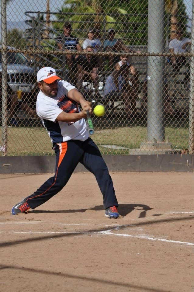 Softball player in white and orange swings at a pitch on a dirt field, with spectators behind a chain-link fence