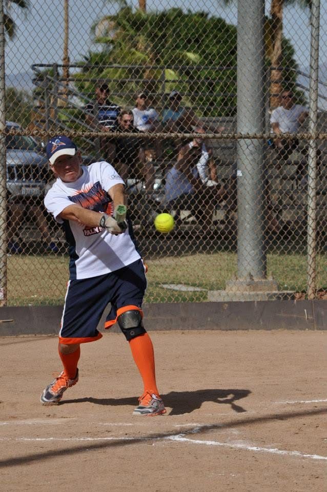 Softball player in white and navy swings at a yellow ball on a dirt field behind a chain-link fence.