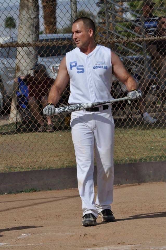 Man in white athletic gear standing on a dirt field, holding a bat behind a chain-link fence.