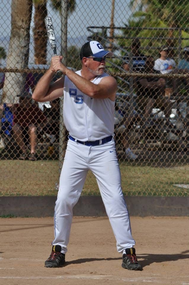Baseball player in white uniform batting in a sandy field, with a chain-link fence and spectators behind him.