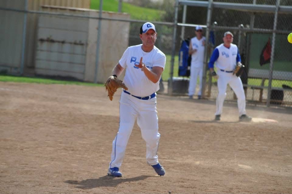 Baseball player in white uniform ready to catch a ball on a dirt field, with teammates near the dugout behind.