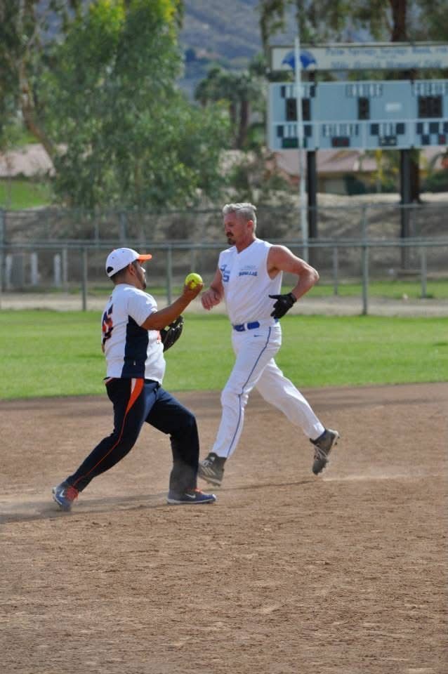 Two softball players high-five on a dirt field, with one holding a yellow bat and a building in the background.