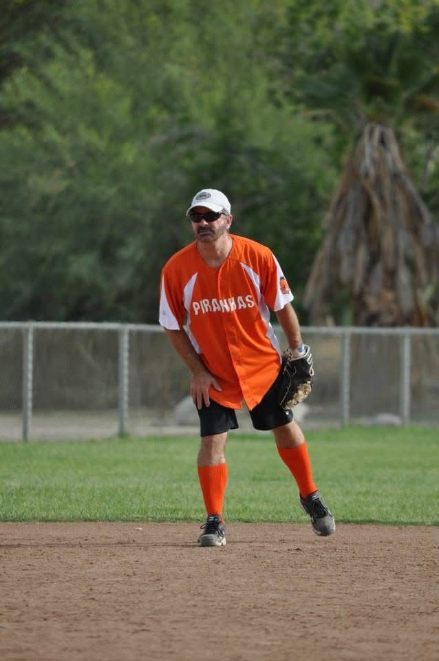 Softball player in orange uniform and cap stands ready on the infield dirt.