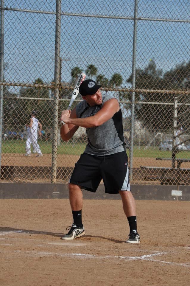 Baseball batter in gray shirt and black shorts at the plate, preparing to swing on a dirt field.