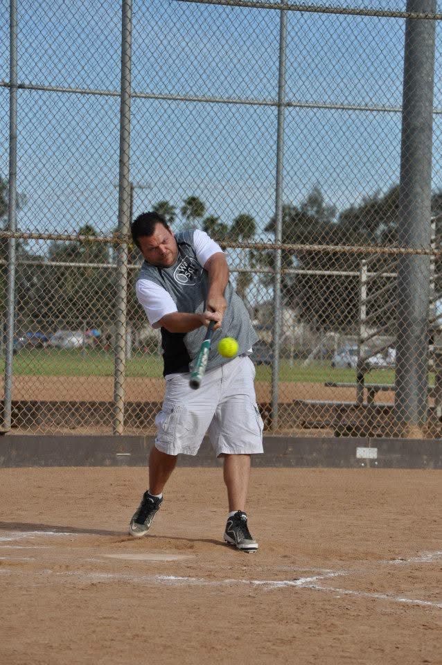 Baseball player swinging a bat at a yellow ball on a dirt field near a chain-link backstop.