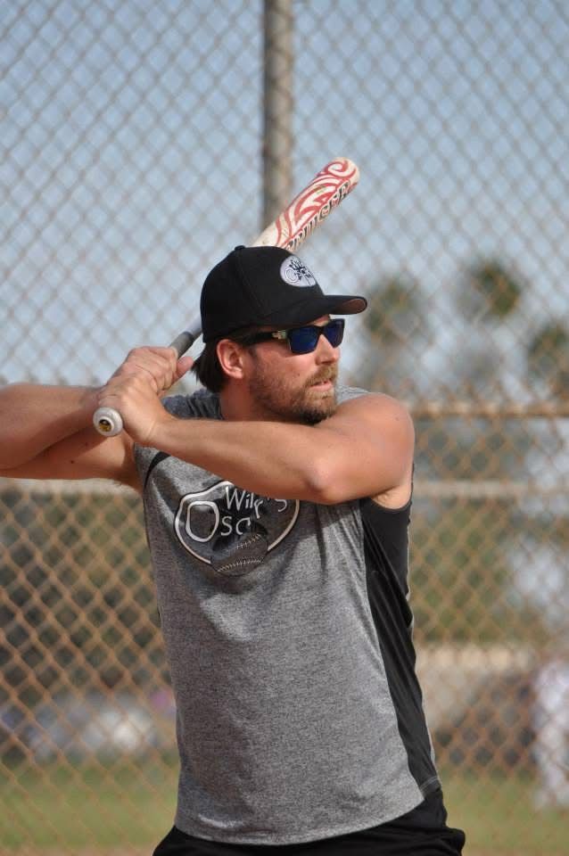 Baseball player in gray shirt and black cap swings a bat on a field, with chain-link fence behind him