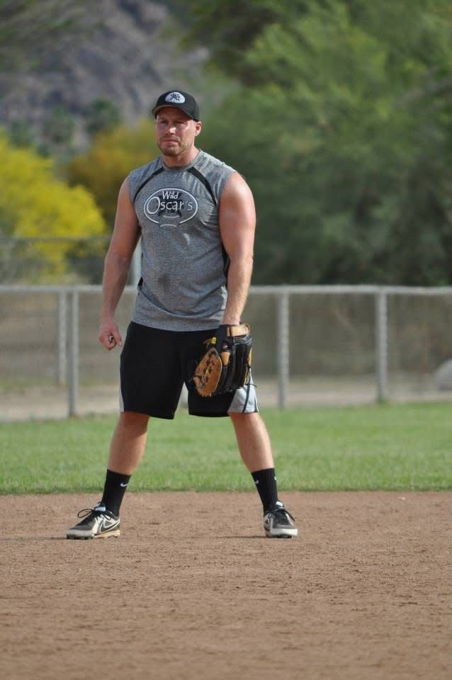 Baseball player standing on a dirt infield, holding a glove, in a gray sleeveless shirt and black shorts.