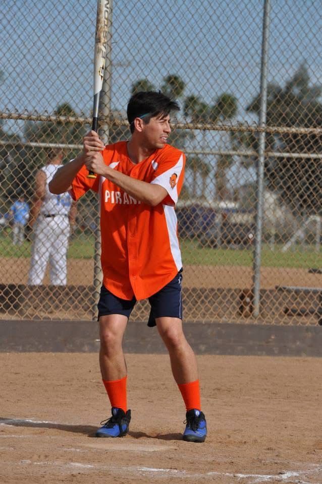Softball player in orange jersey stands at bat on a dirt field behind a chain-link fence.