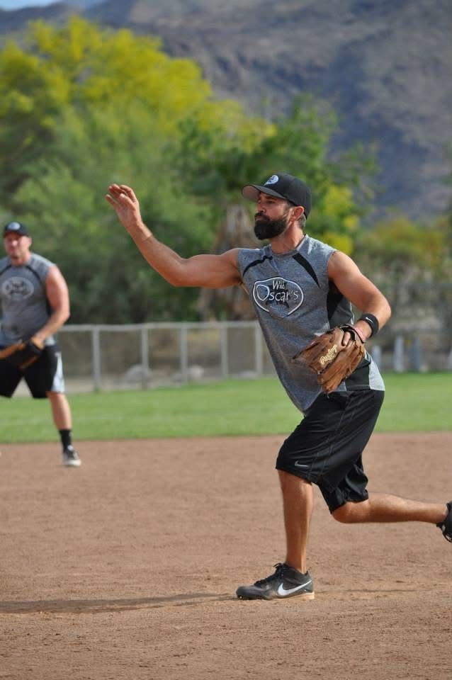 Softball player in gray shirt and black cap throws a ball on a dirt field with a mountain backdrop.
