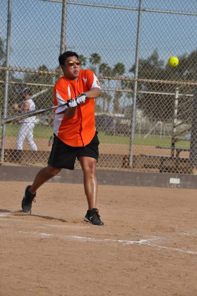 Softball batter in orange shirt swings at a yellow ball on a dirt field.