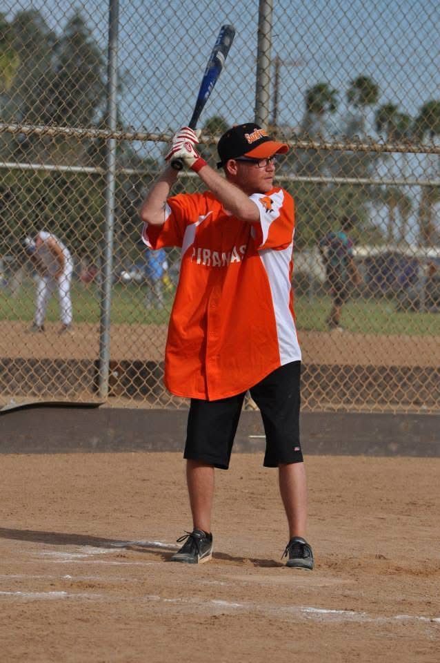 Softball batter in orange jersey stands ready at home plate with bat raised on a field.