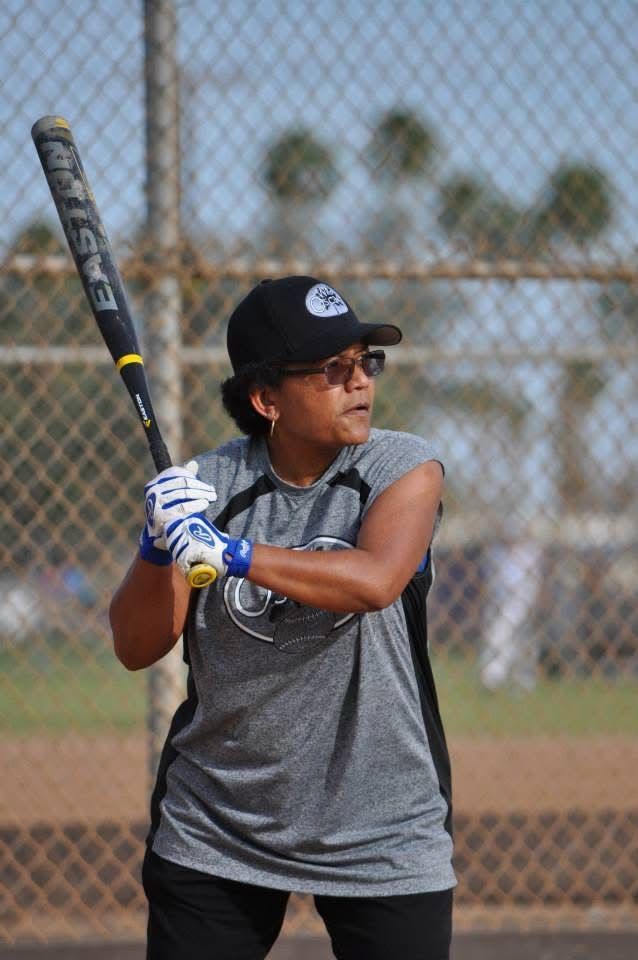 Softball player in gray shirt and black cap holding a bat in batting stance on a field