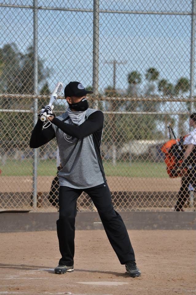 Baseball batter in black gear stands ready at the plate on a dirt field with a chain-link fence behind him