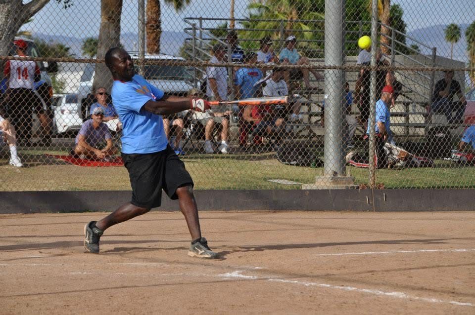 Softball player in blue shirt swings at a pitch on a dirt field with spectators behind the fence