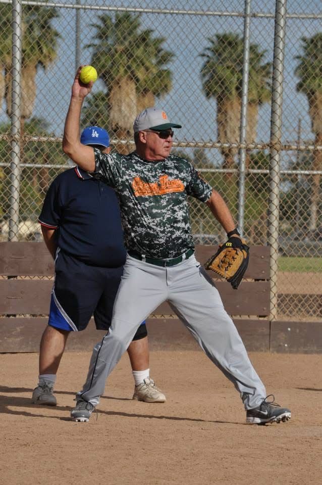 Softball player in camo jersey and gray pants prepares to throw on a dirt field behind a chain-link fence.