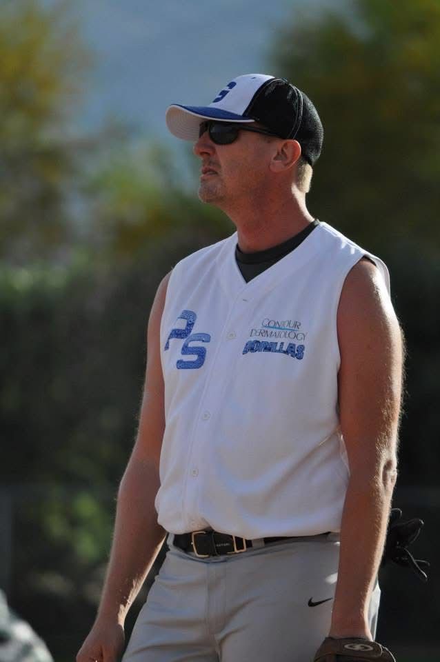 Man in white sleeveless shirt and cap standing outdoors in sunglasses under bright sun