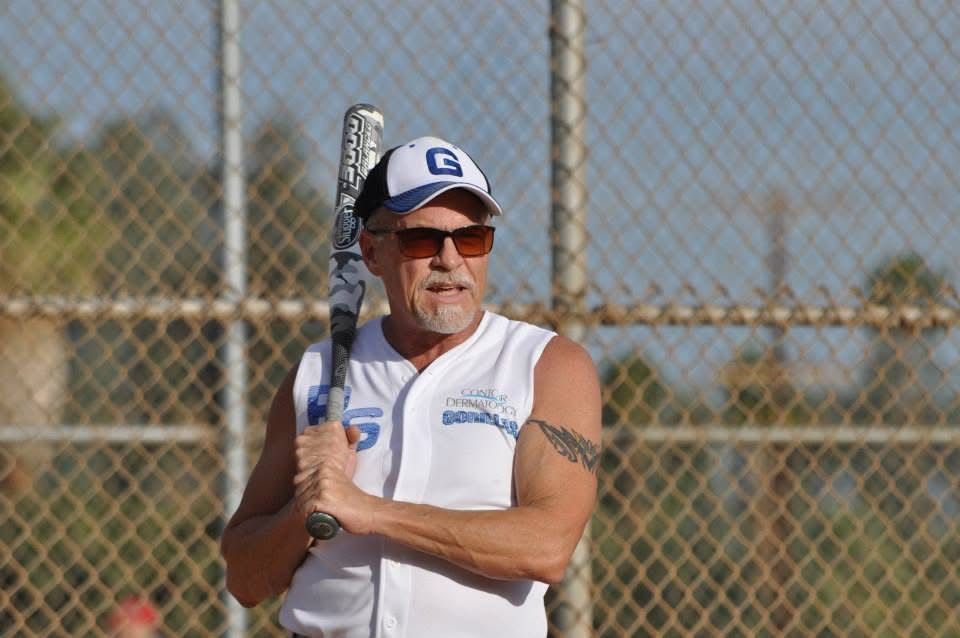 Man holding a baseball bat on a field, wearing a white jersey and cap near a chain-link fence.
