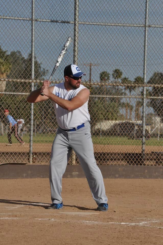 Baseball batter in gray uniform holding a bat at the plate, with a chain-link fence and field behind him.